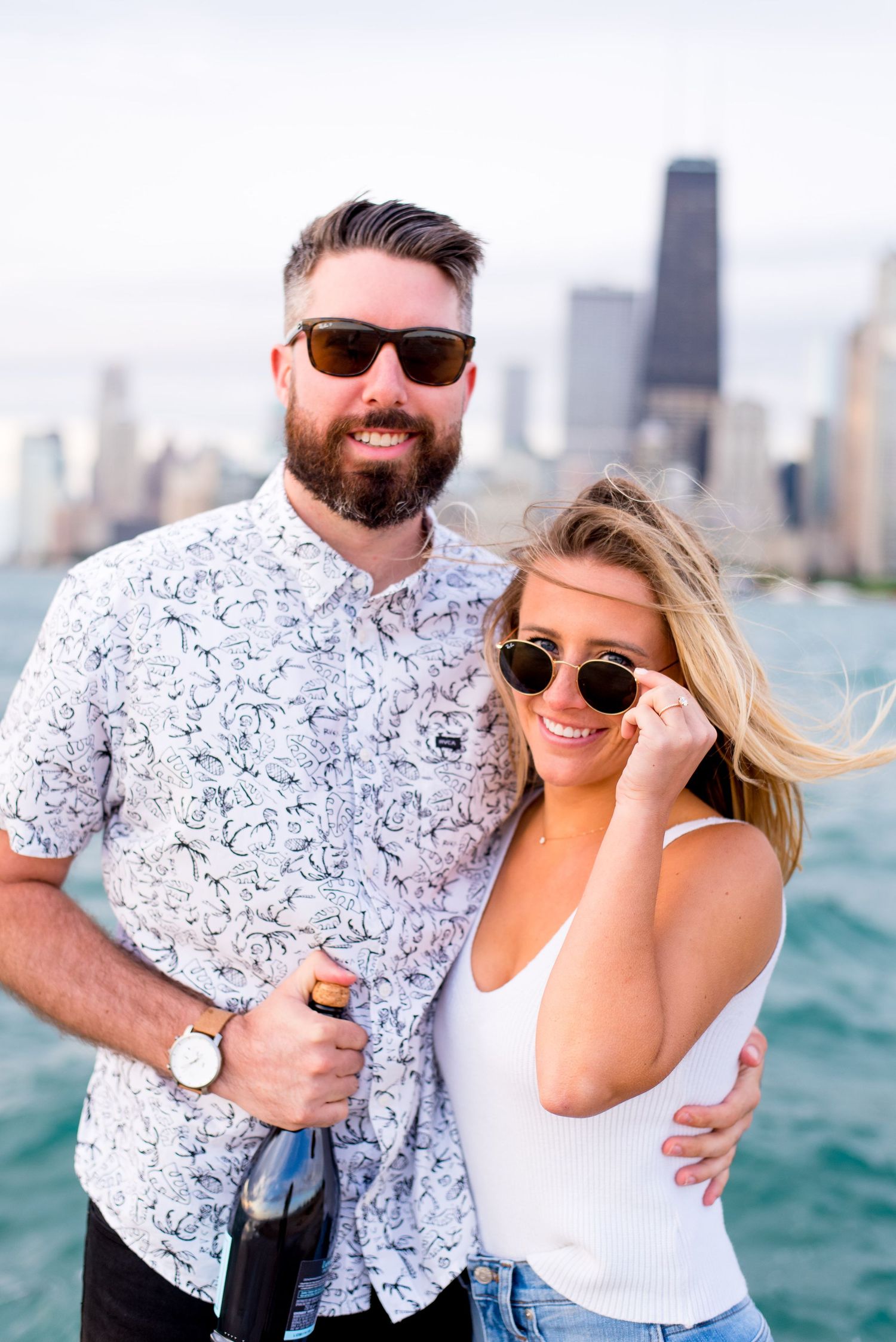 engaged man and woman wear sunglasses and smile in the wind and hold champagne at North Avenue Beach Chicago
