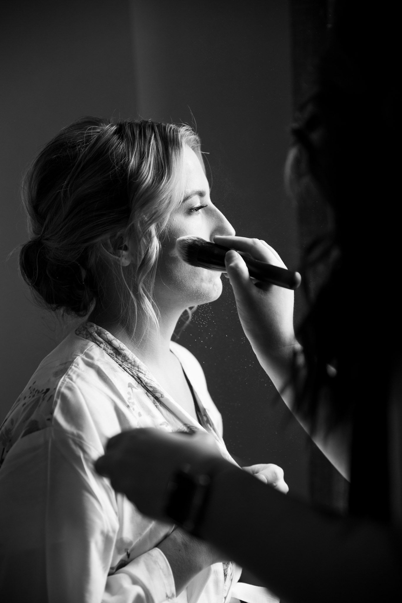 black and white image of bride getting blush on her face and makeup fallout in the light