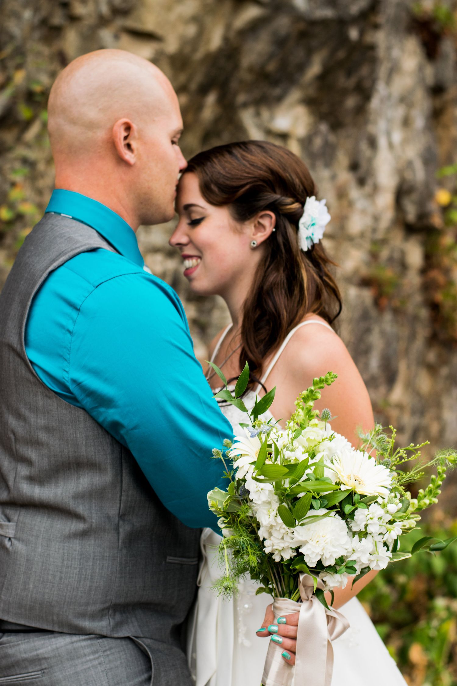 Tender moment between newlyweds with white floral bouquet against rocky backdrop.