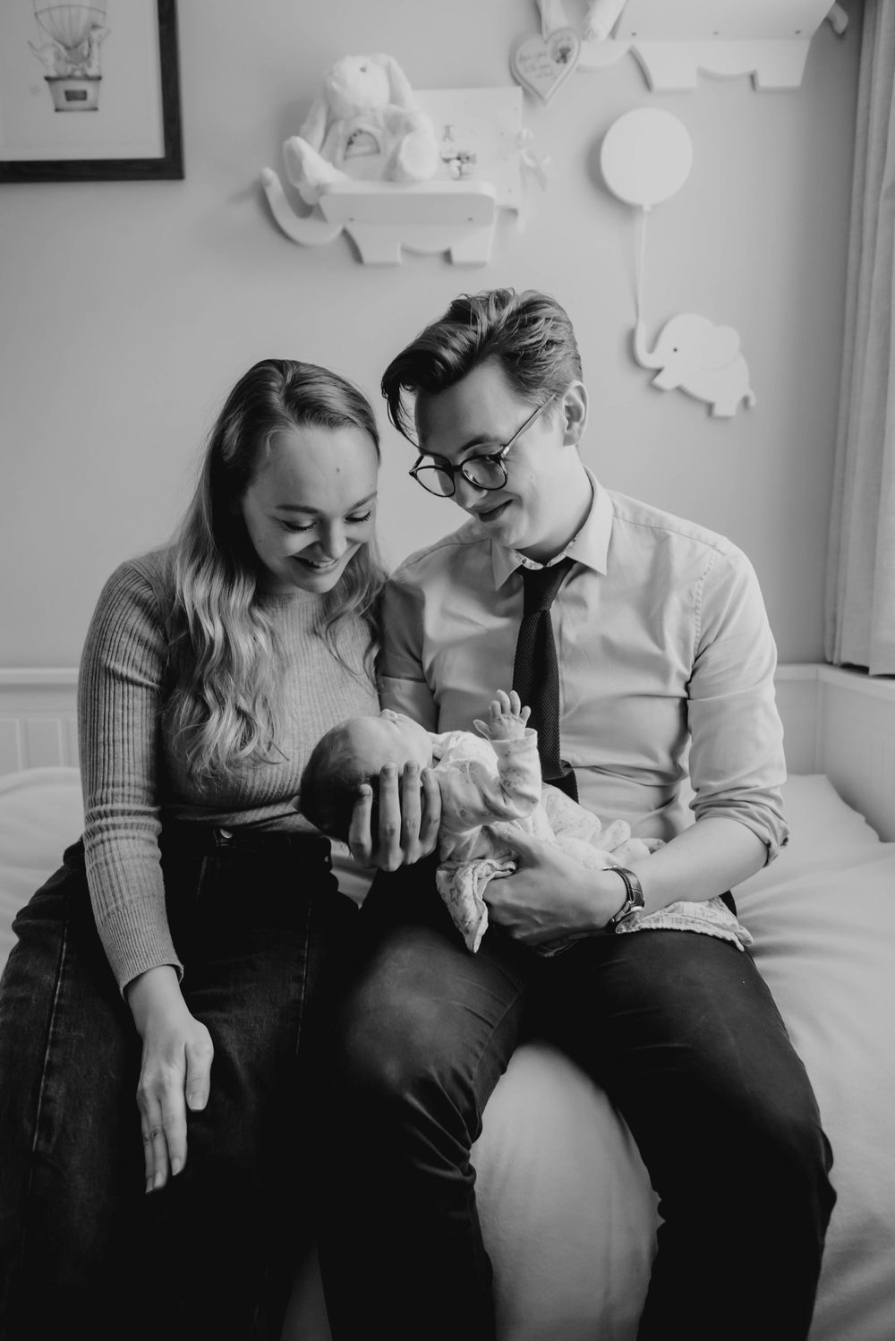 A couple sits together on a couch looking lovingly at their newborn baby in a nursery setting.