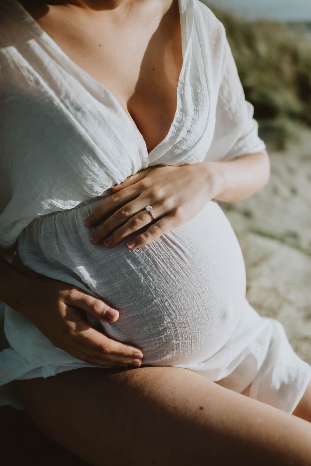 Close-up of hands cradling pregnant belly in white lace dress against natural backdrop.