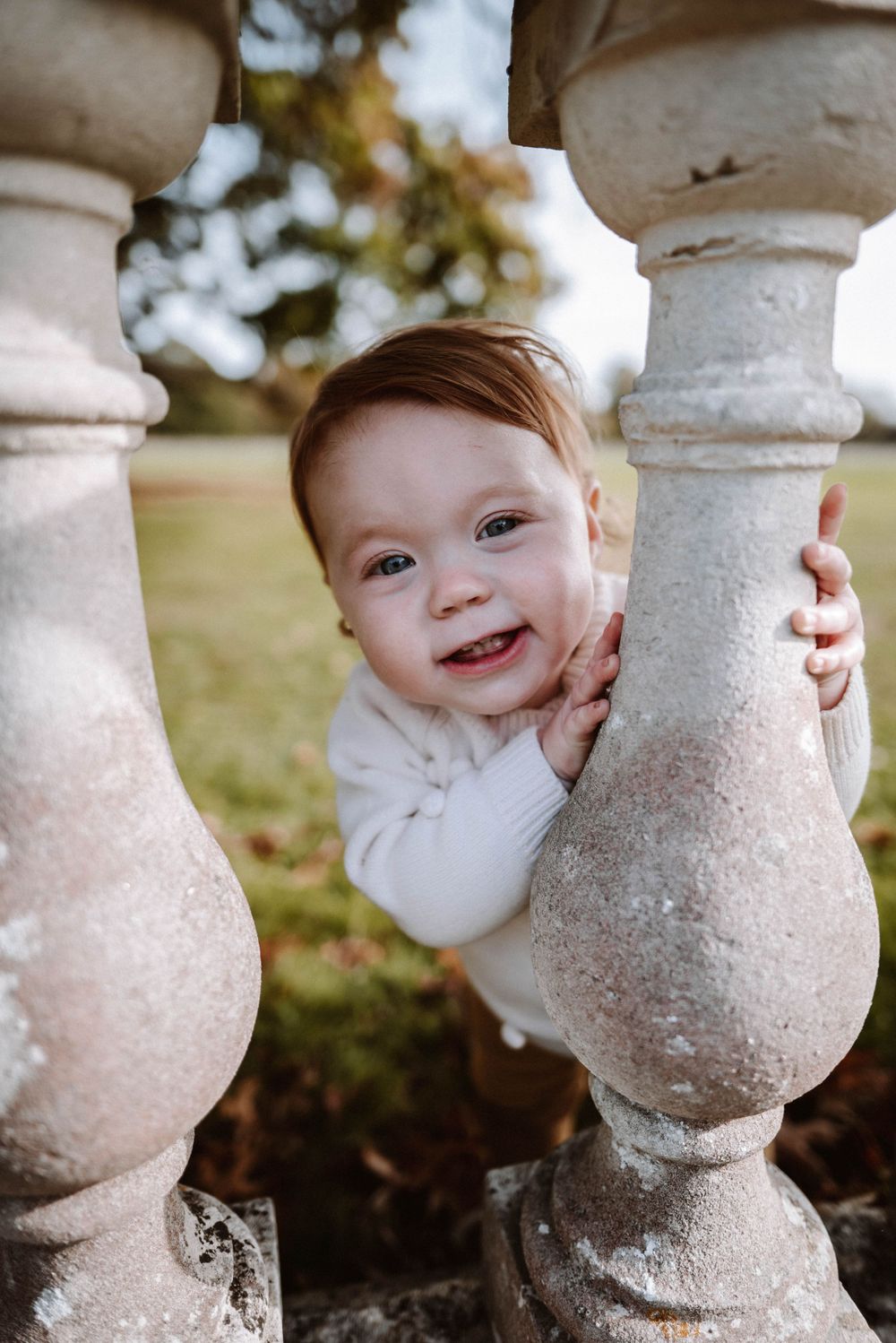 A playful child peeks through stone balusters with a bright smile outdoors.