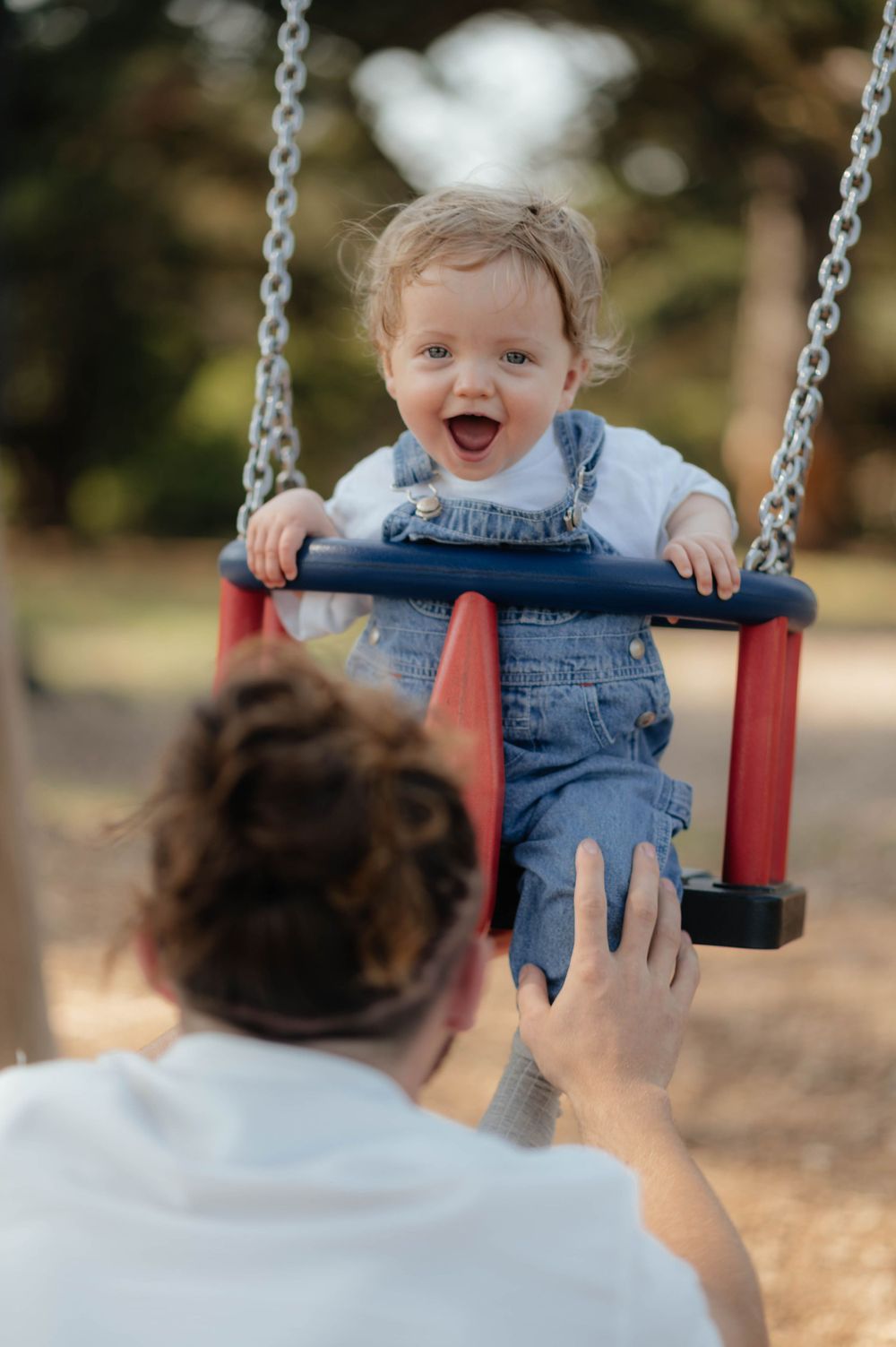 A joyful baby in denim overalls enjoys swinging at a playground.