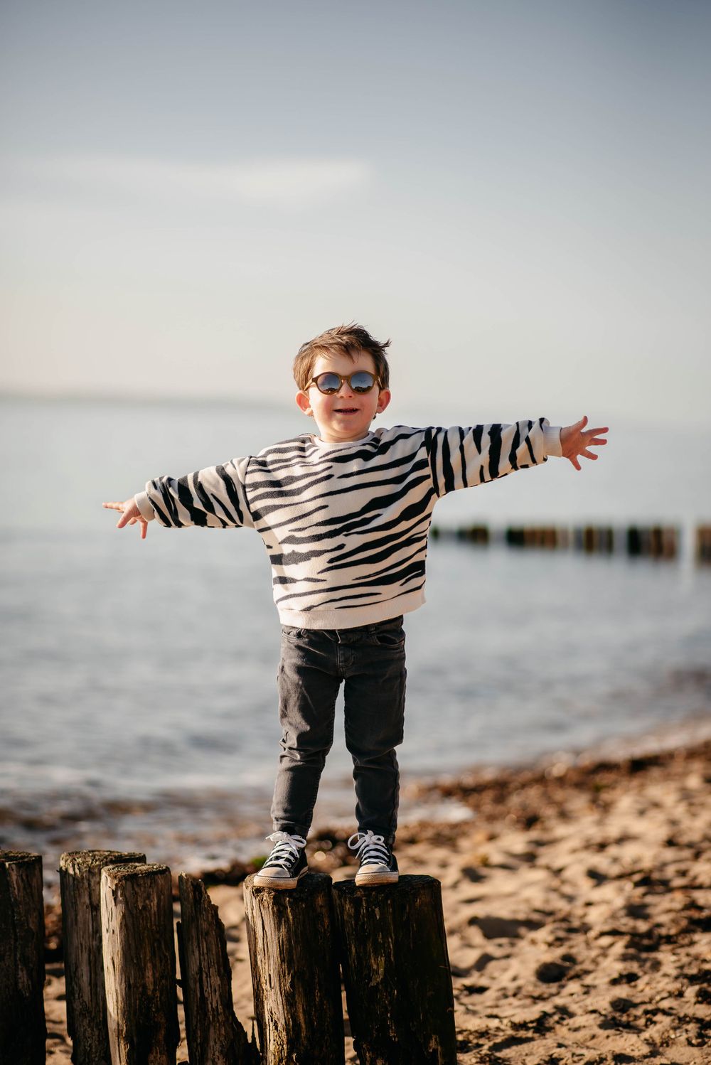Child in striped sweater stands with arms outstretched on beach post at sunset.