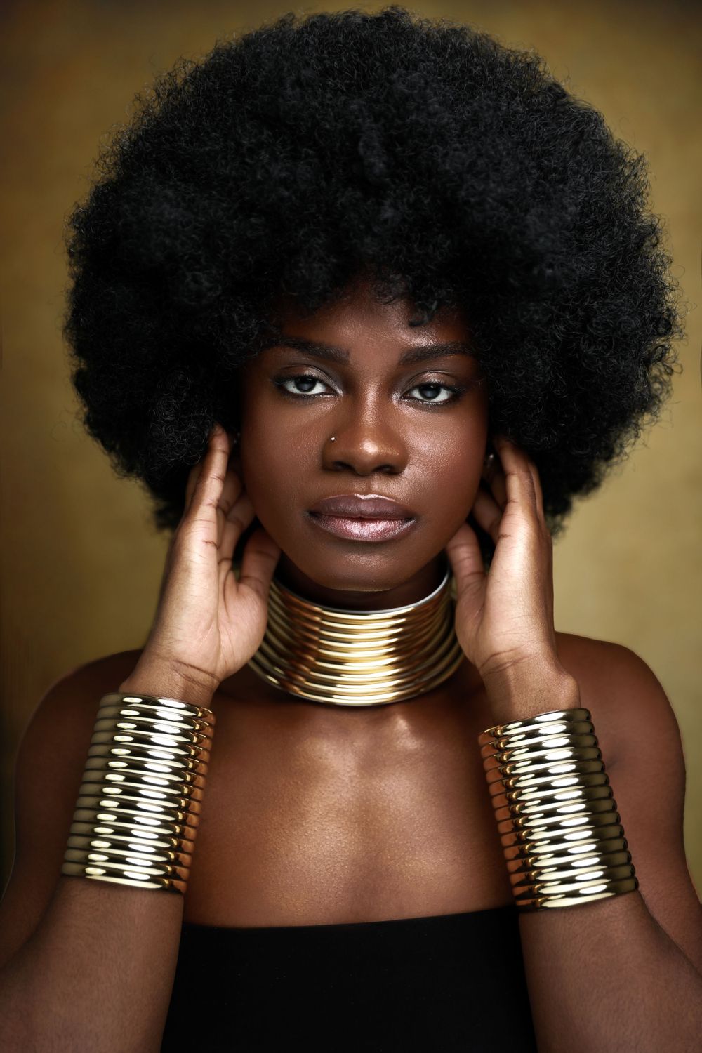 Portrait of woman with natural afro wearing gold neck and arm cuffs against golden background.