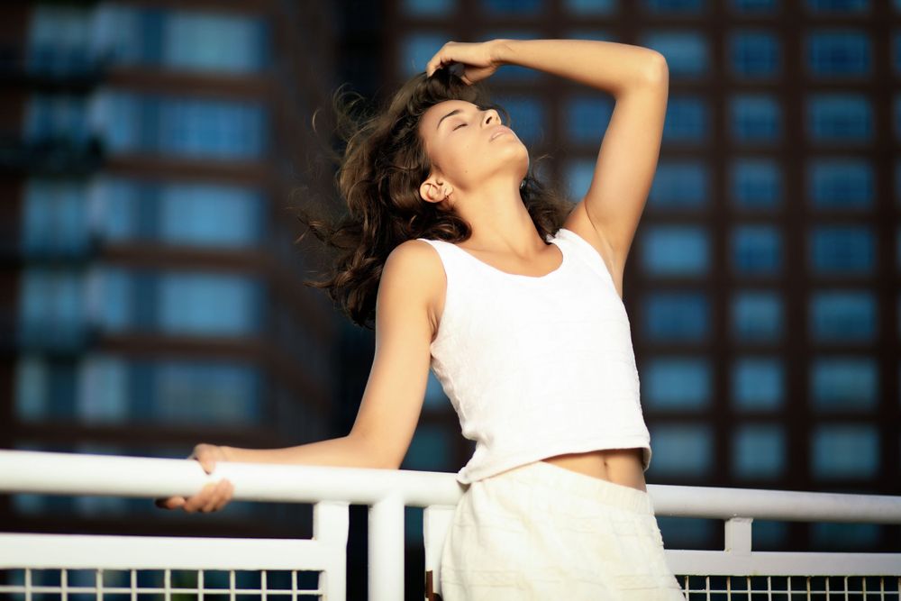 Woman in white tank top and beige pants posing expressively against urban railing with city background.