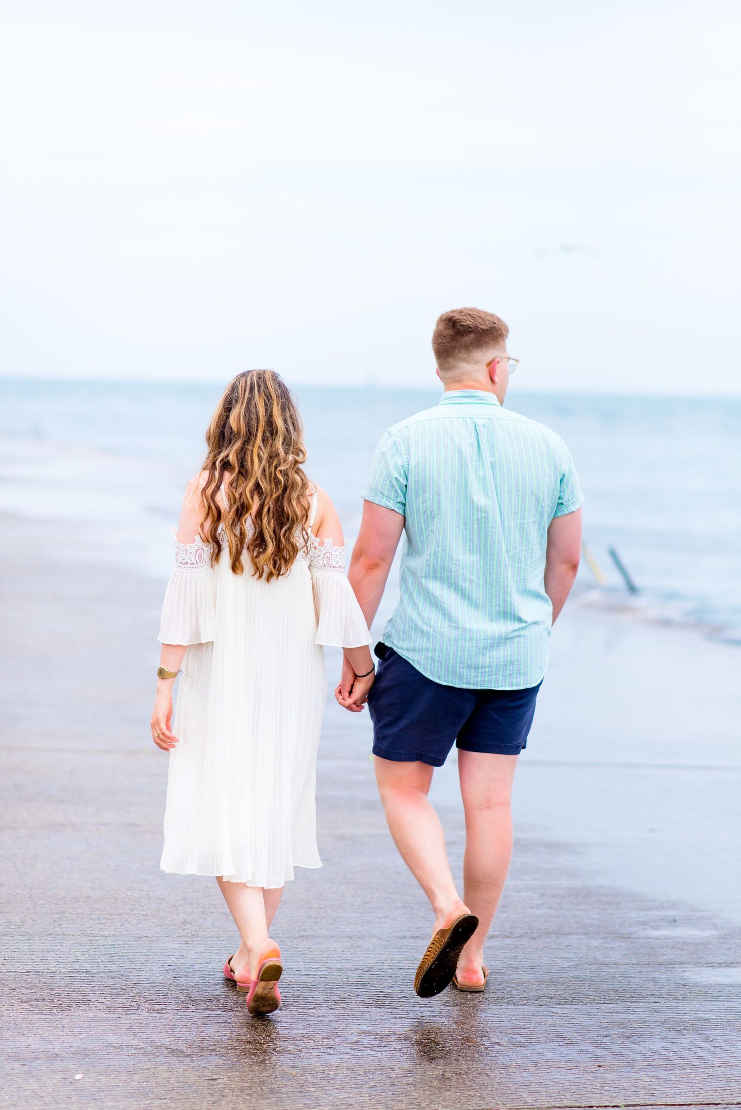 young man in blue shirt and fiancee in white dress walk along Montrose Harbor holding hands at sunrise