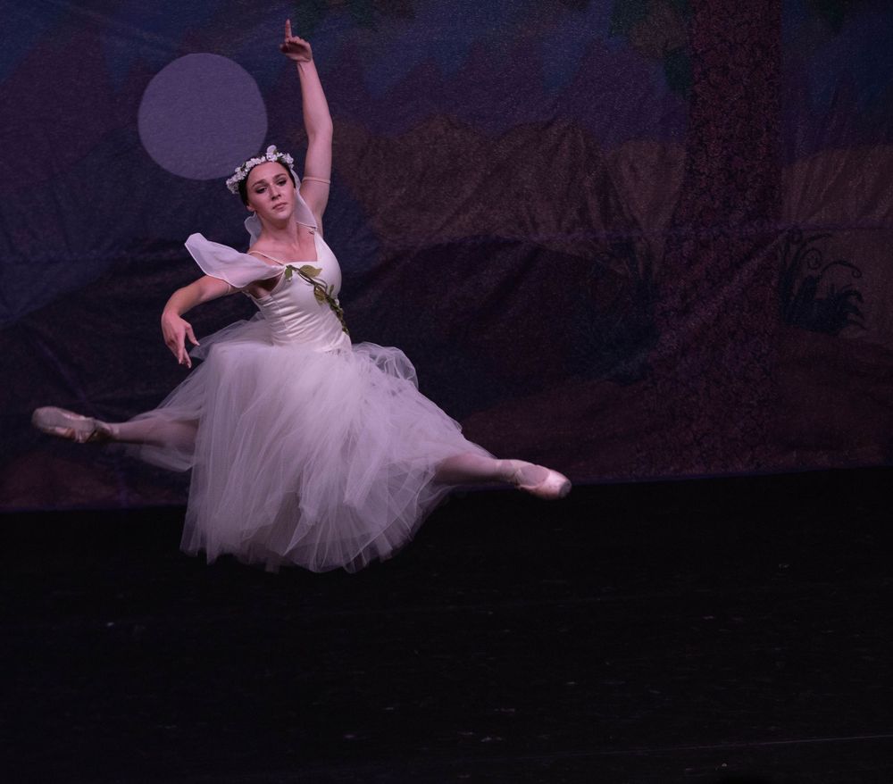 Ballet dancer in white tutu performing graceful leap on dark stage.