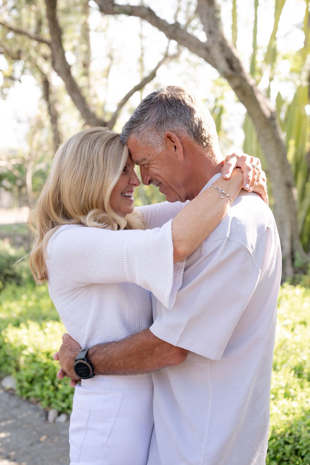 A loving mature couple embraces while wearing white outfits in a sunny garden setting.