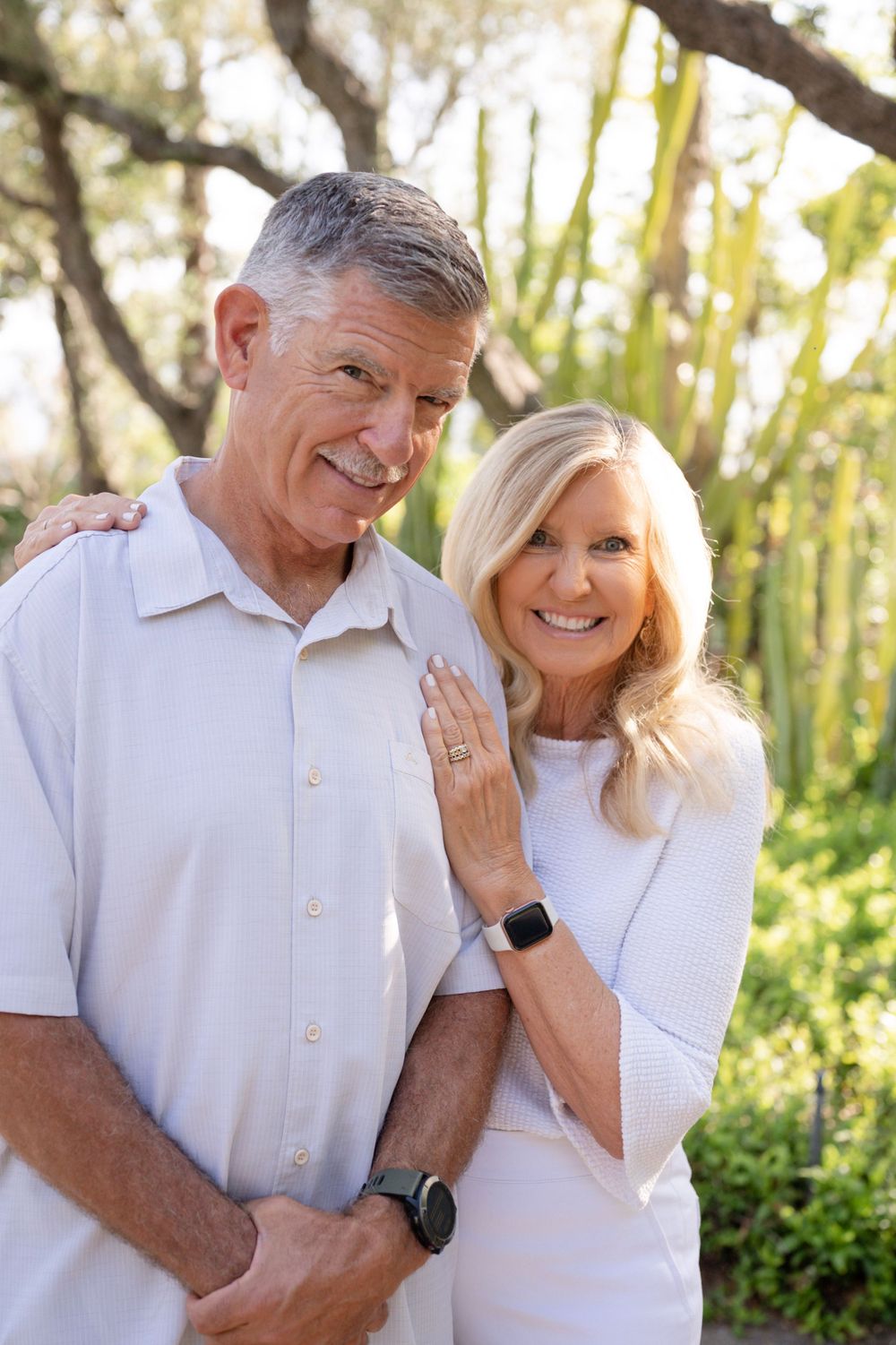 Happy couple dressed in white poses together against a background of trees and greenery.