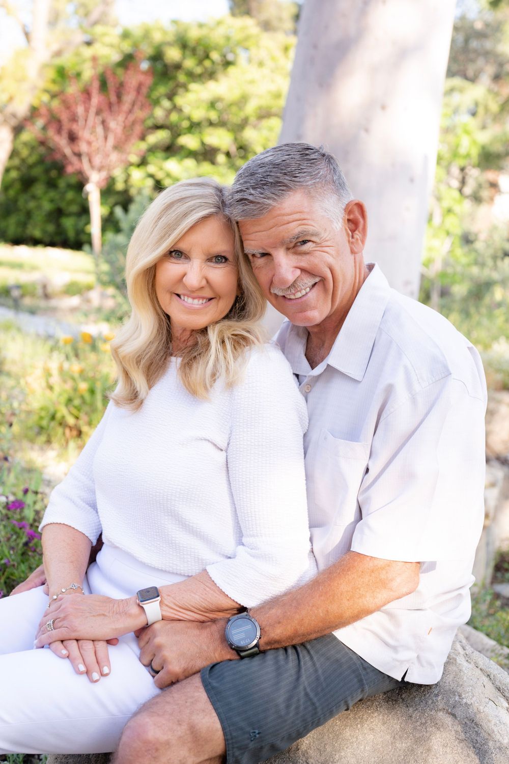Couple sitting close together on a rock in a garden setting wearing coordinated white outfits.