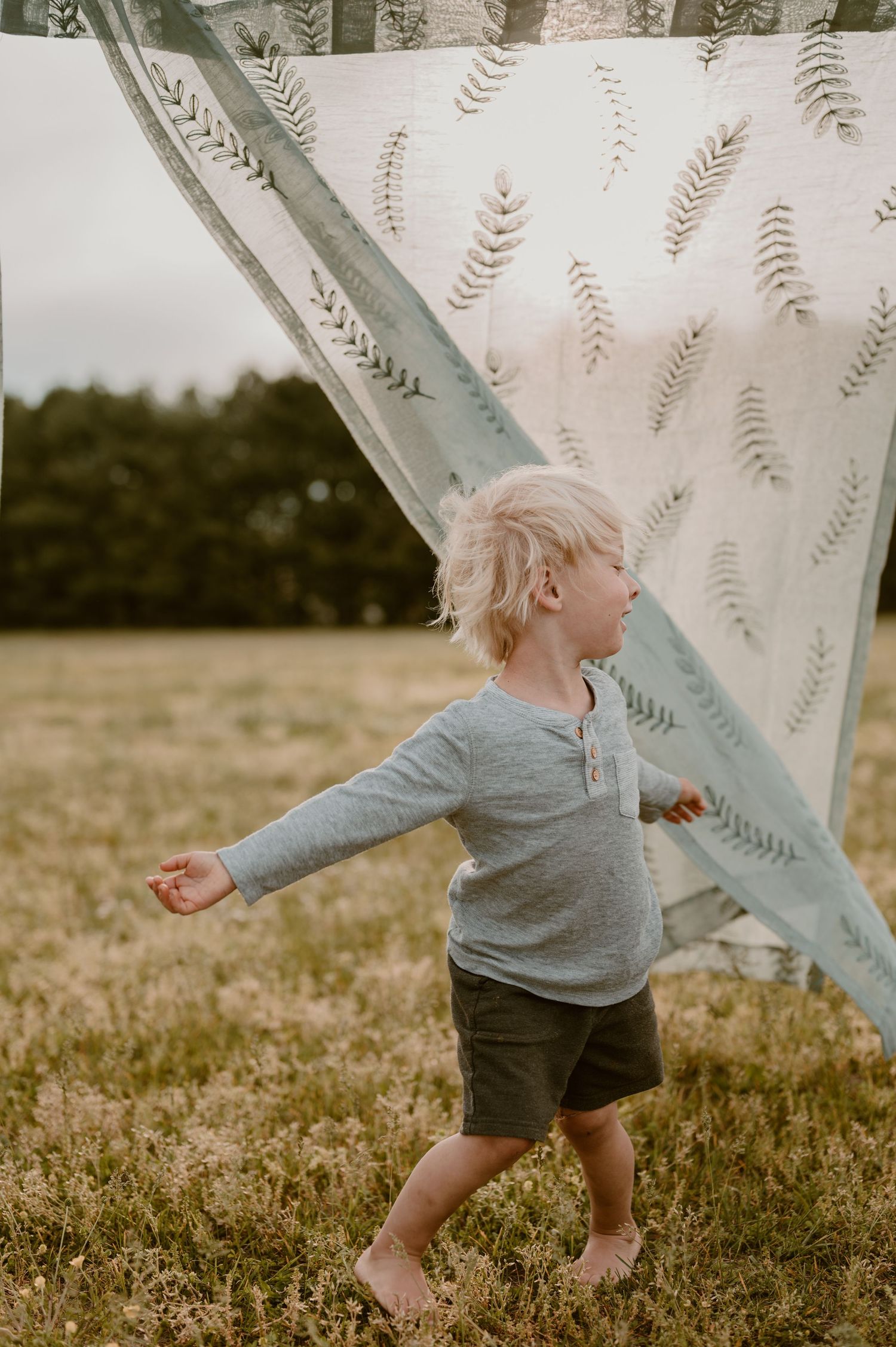A small child in grey plays by a billowing white curtain with botanical patterns in a sunlit field. photo by Samantha Campbell imagery