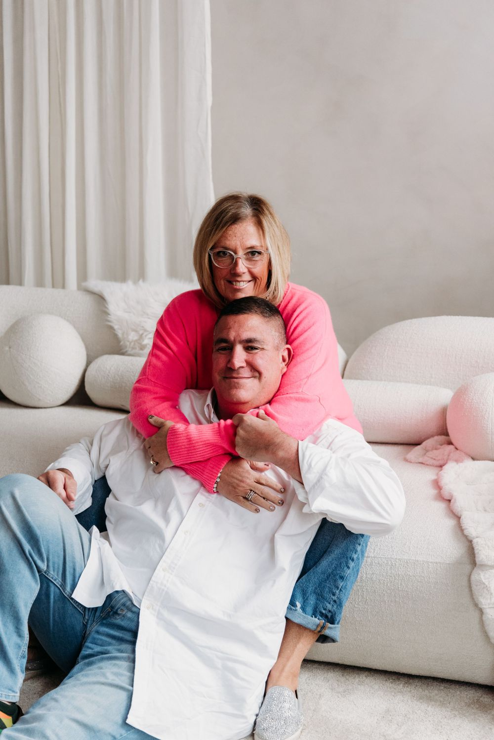 Playful indoor portrait of couple embracing on white couch with pink accents.