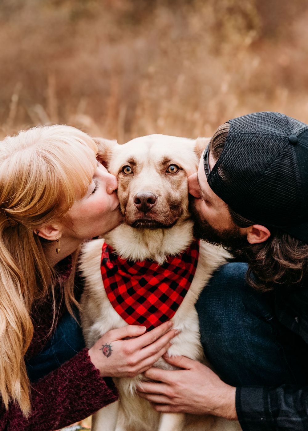 Dog wearing buffalo plaid bandana gets kisses from both sides in autumn setting.