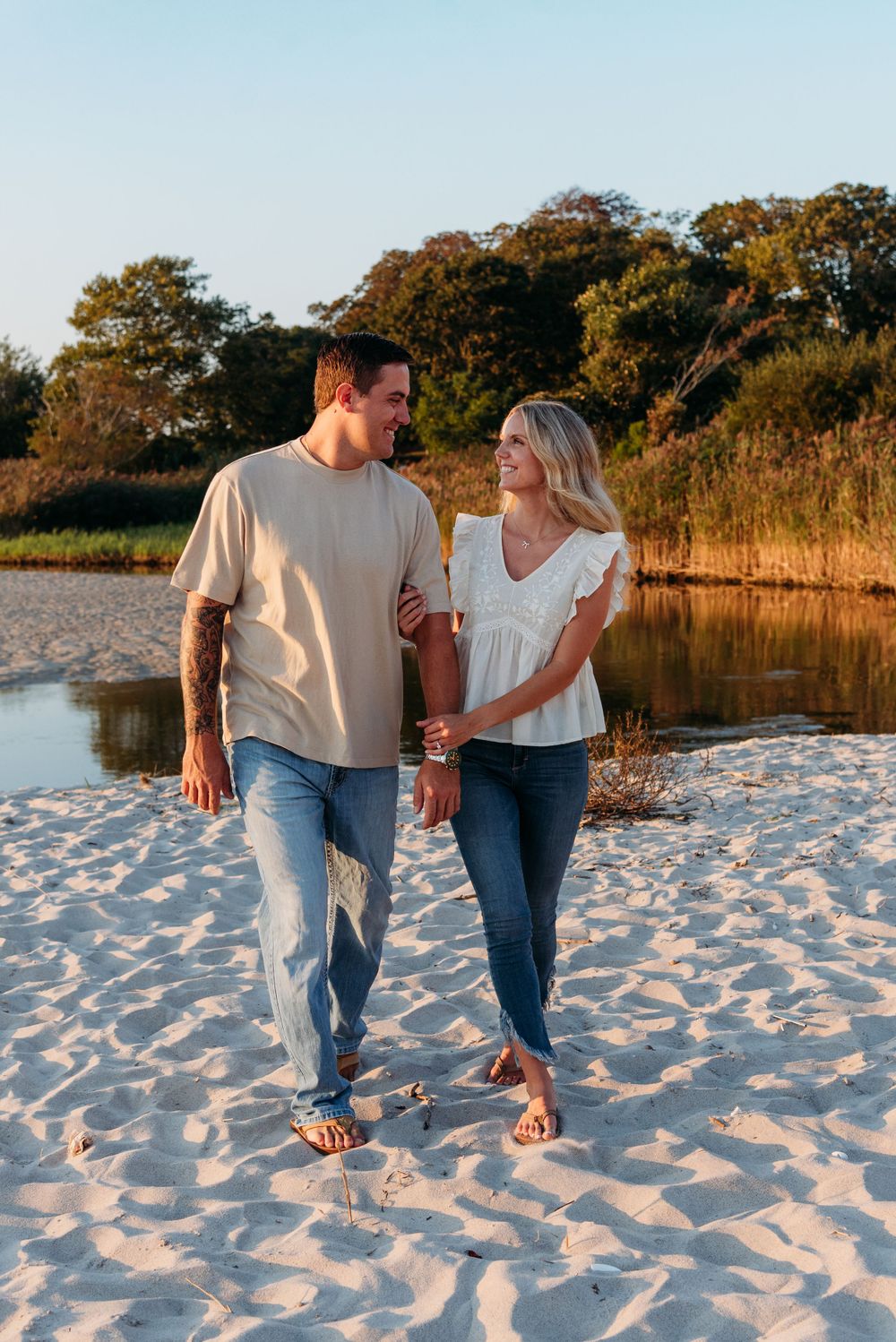 A couple takes a romantic sunset stroll along a sandy beach with trees and marsh in the background.