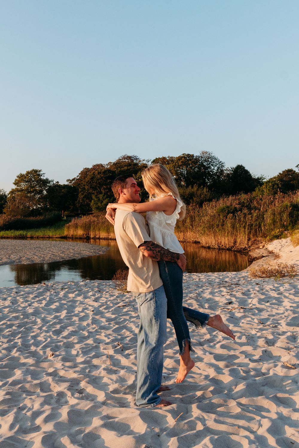 A romantic embrace on the beach with sunlit water and trees in the background.