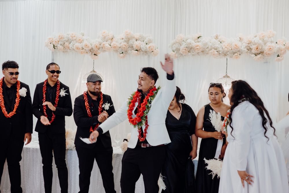 Wedding party members in black attire and traditional leis celebrate against a white backdrop with pampas grass decor.