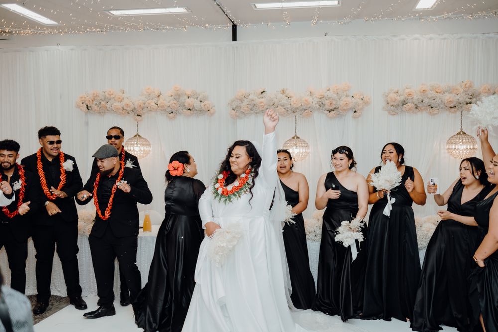 Wedding celebration with party members in black attire dancing against white floral backdrop.