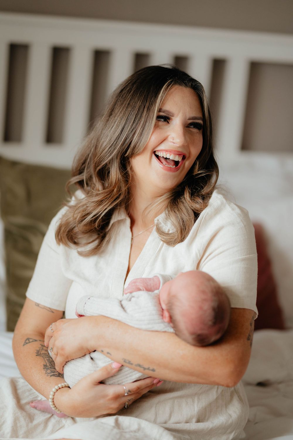 A joyful mother in white clothing holds and smiles while cradling a newborn baby on a couch.