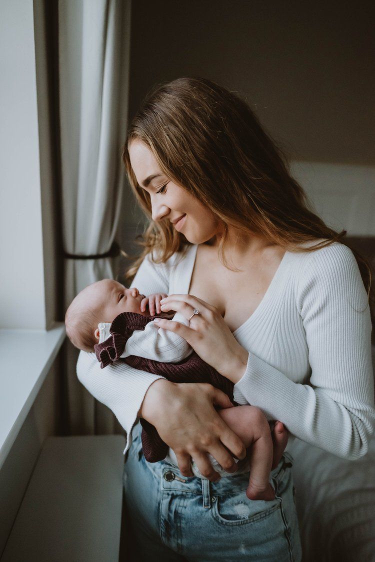 A mother in a white sweater and jeans holds her newborn baby tenderly by a window in soft natural light.