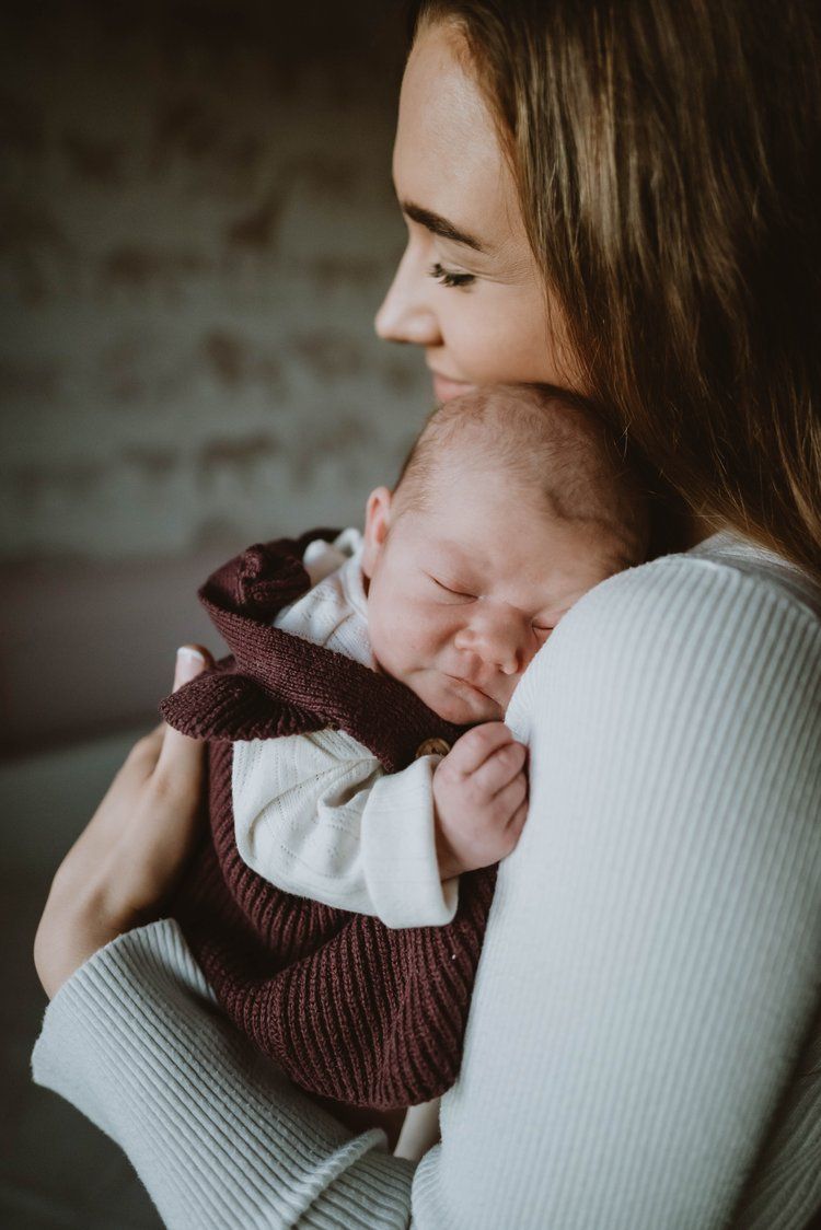 A sleeping newborn in a brown knit outfit rests peacefully against a woman wearing a light gray sweater.