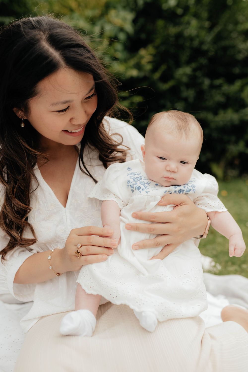 A parent in a white dress holds a baby wearing a white christening gown while sitting outdoors.