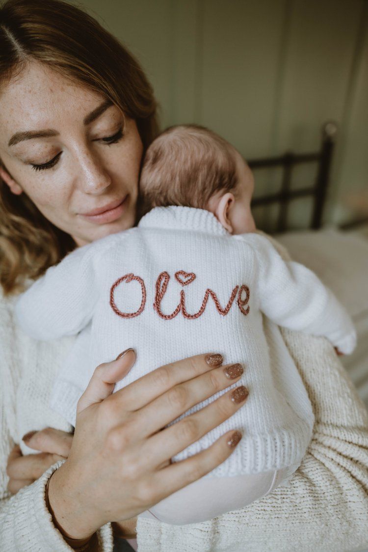 Baby wearing white knit sweater with 'Olive' embroidered on back being held against a cream sweater.