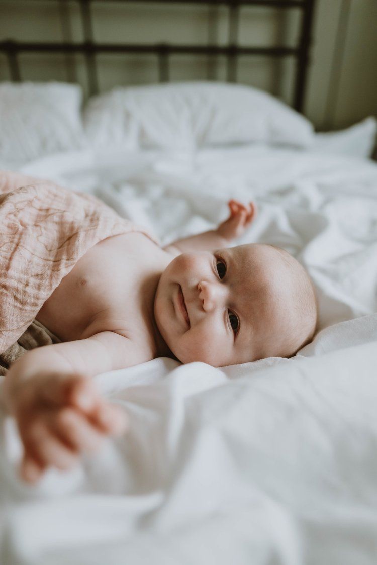 A sweet baby lies on crisp white bedding, wearing a peach-colored outfit while smiling and reaching out.