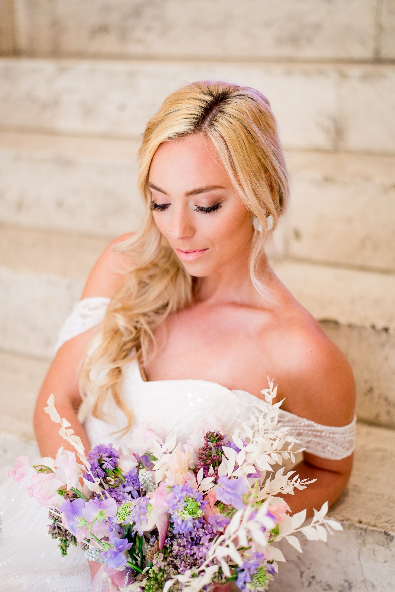 blonde bride with messy braid sitting on marble steps looking at pink purple white wedding bouquet