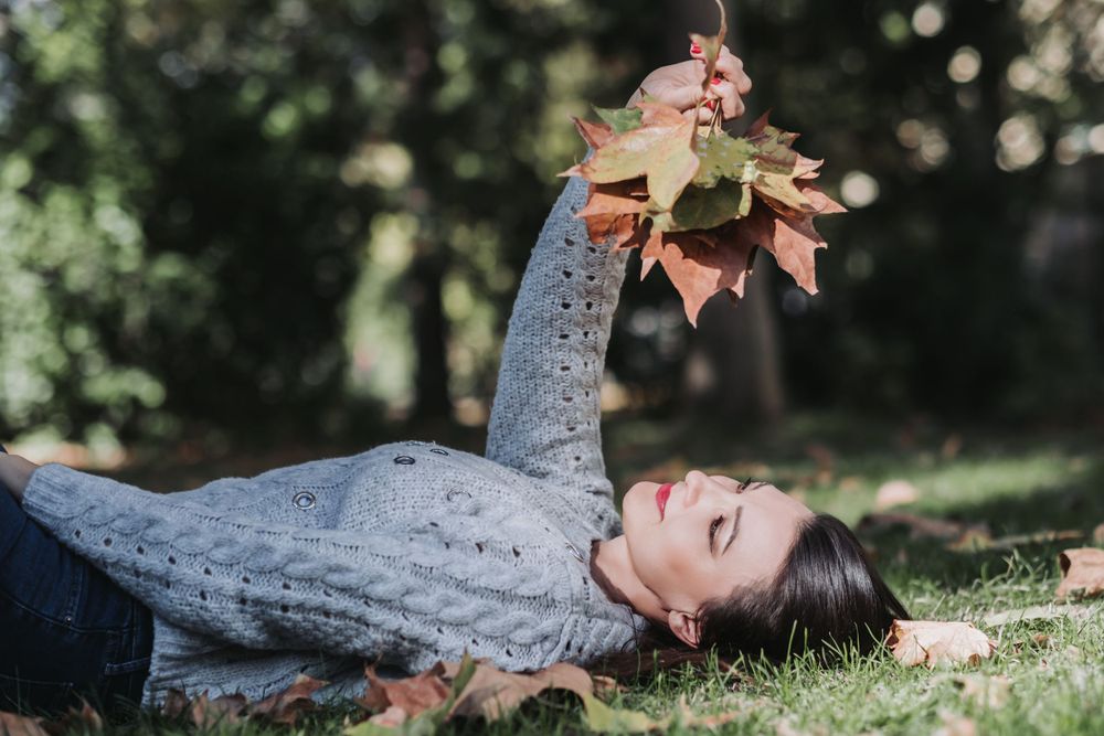 Person in gray sweater lies on ground holding autumn leaves while enjoying nature.