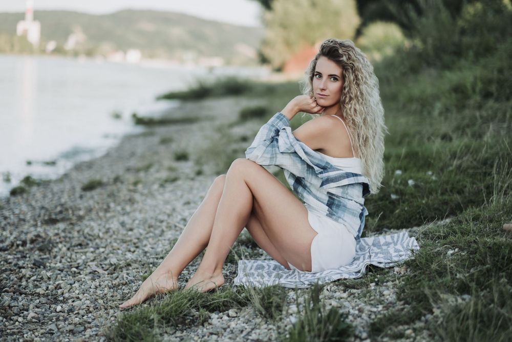 Relaxed pose on a rocky beach wearing denim and plaid against a waterfront backdrop.