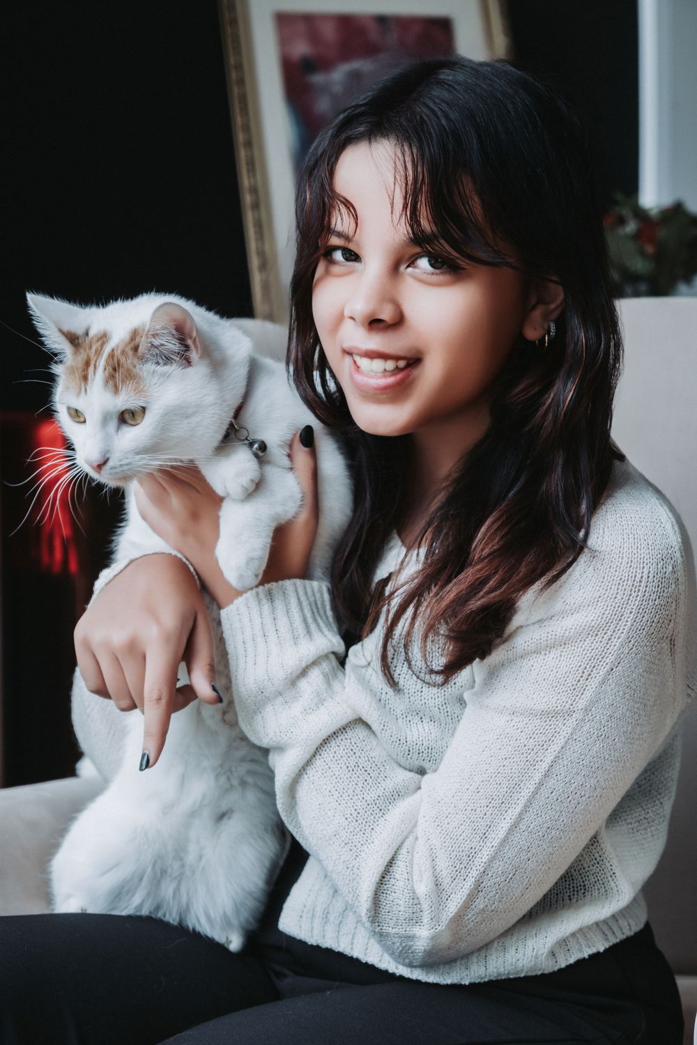 Person in white sweater holds adorable white cat with red collar in indoor setting.