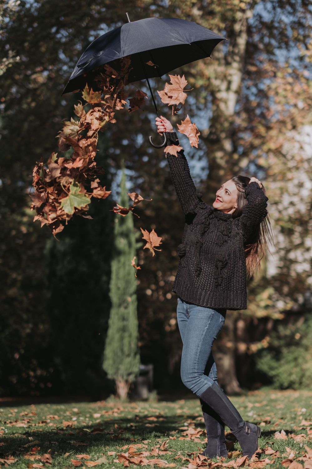 Person in knit sweater playfully tosses autumn leaves while holding black umbrella in park setting.