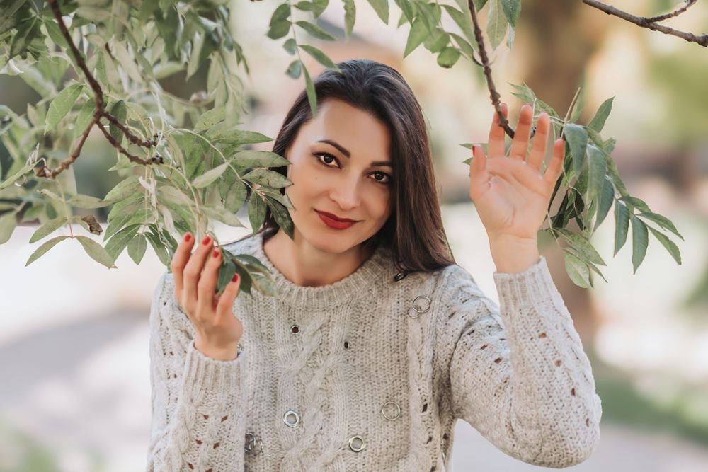 Woman in cozy white knit sweater poses playfully among tree branches in outdoor setting.