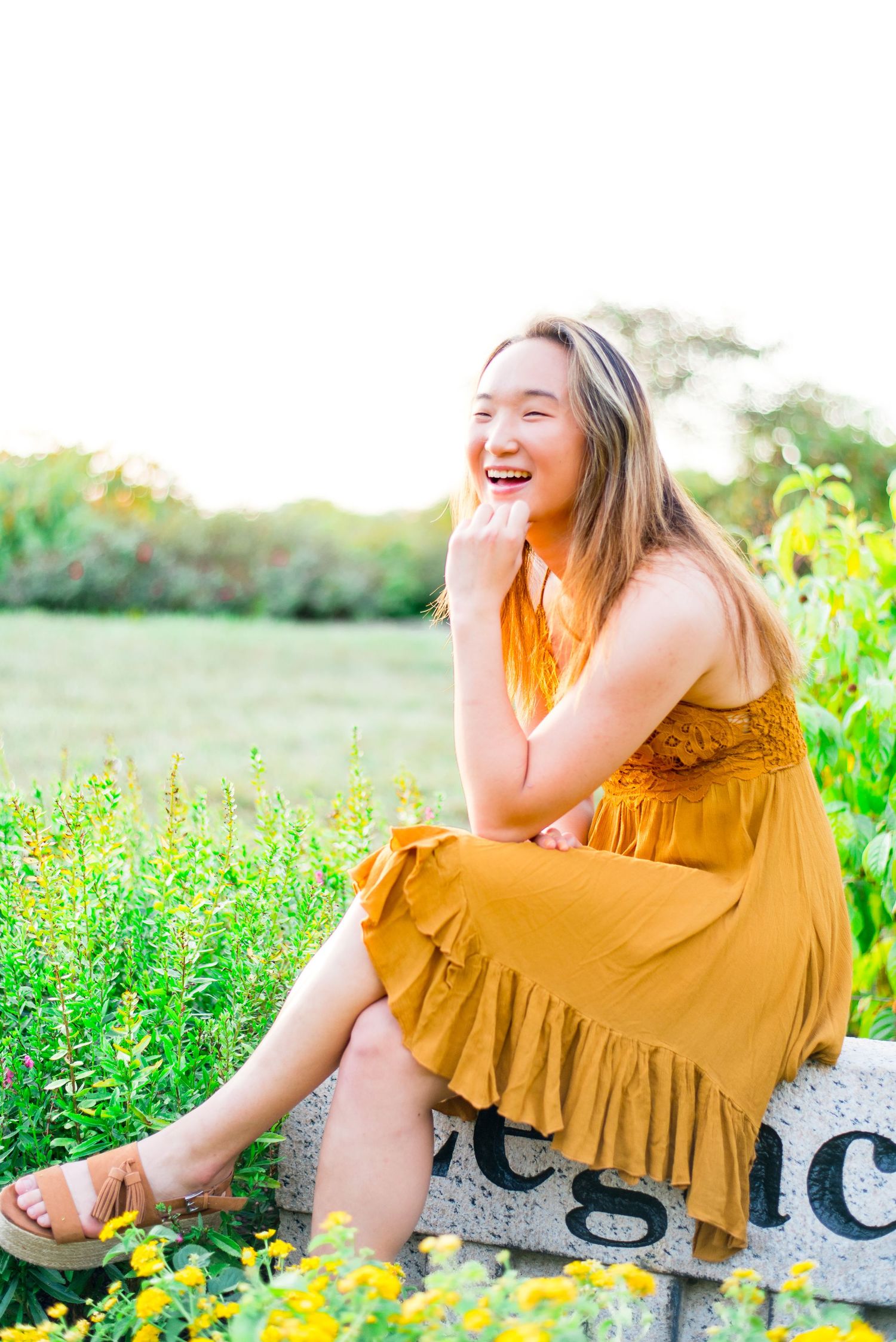 young woman in yellow dress sitting on a stone with knees crossed laughing