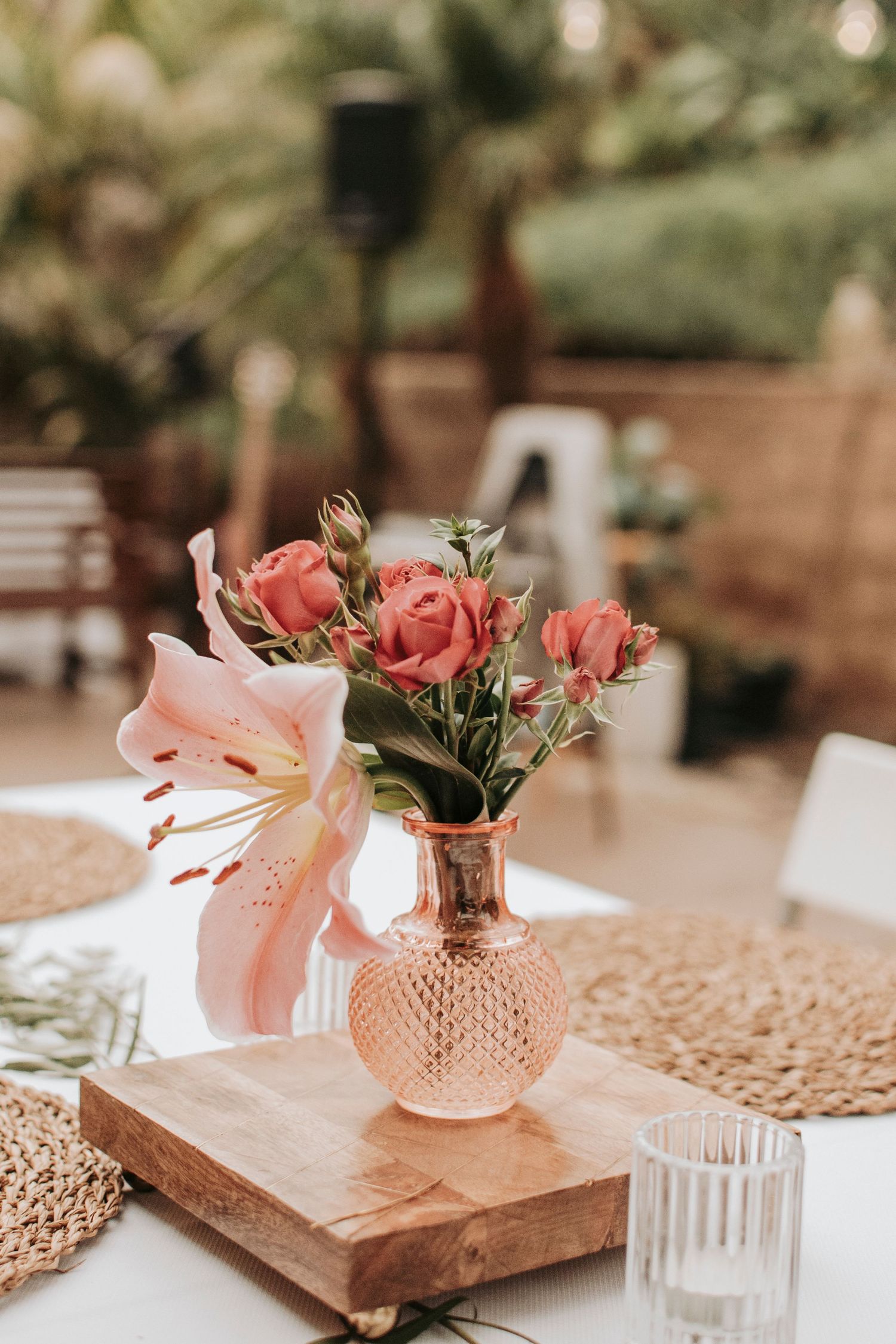 Pink roses and lilies arranged in a vintage-style glass vase on a wooden board with table decor.