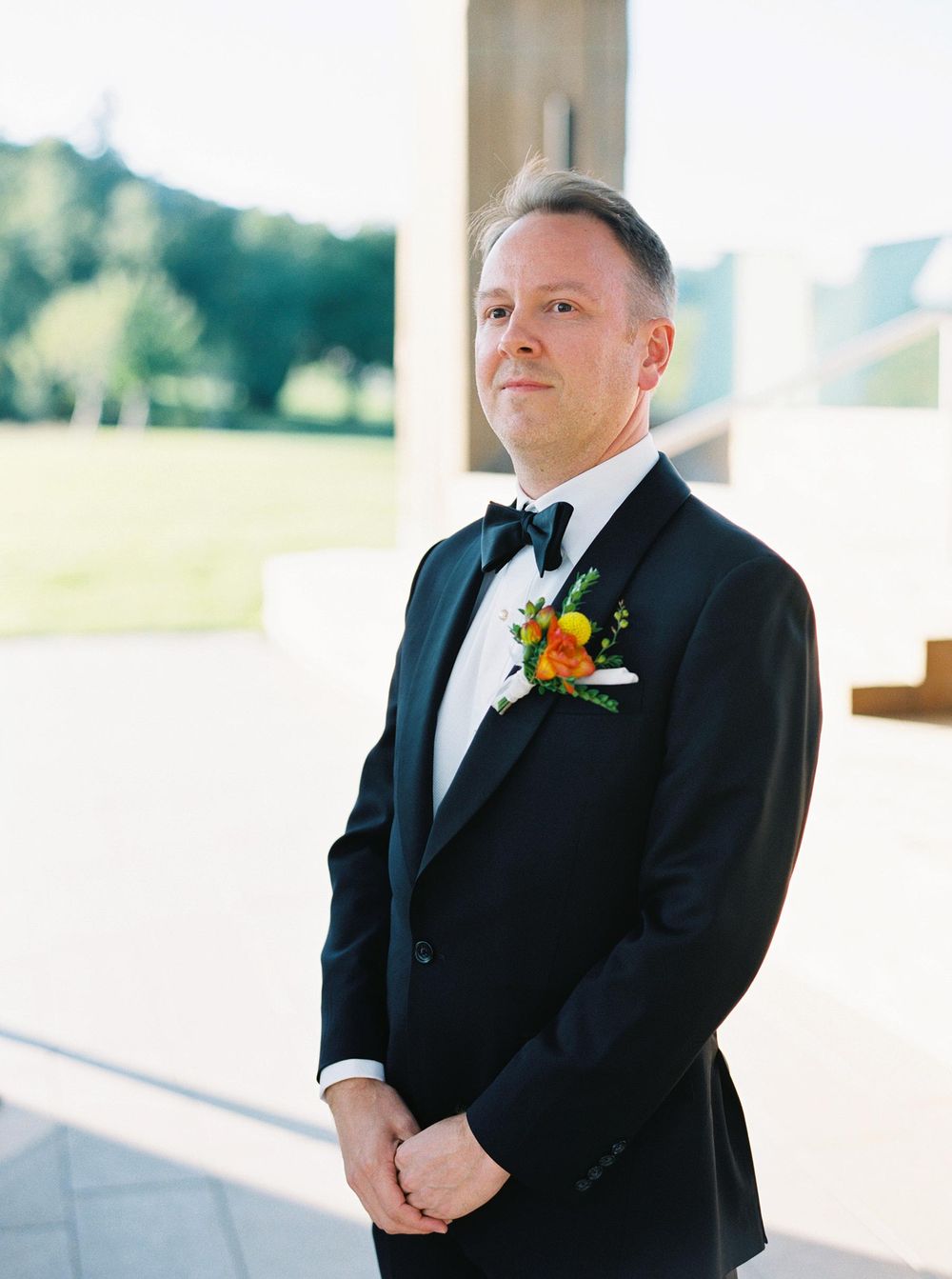 A groom dressed in a tuxedo stands in front of a building, ready to welcome his bride on their wedding day.