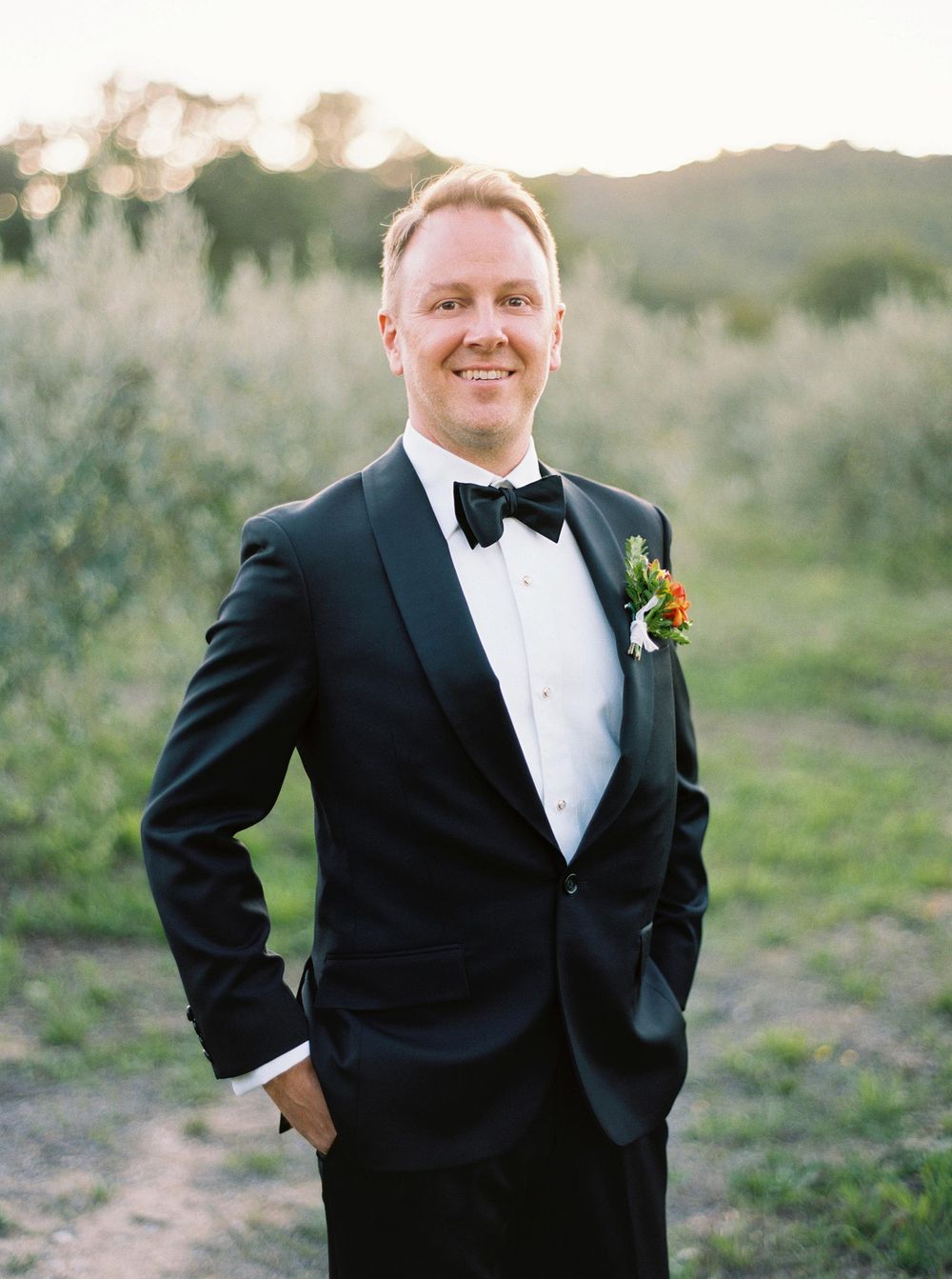 A groom in Provence stands in an olive grove, elegantly posing for a photo in his tuxedo.