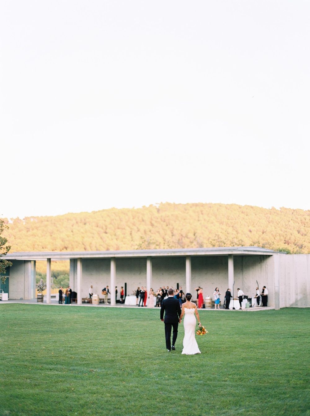 A newlywed couple strolling through lush green grass to their wedding cocktail in Château La Coste.