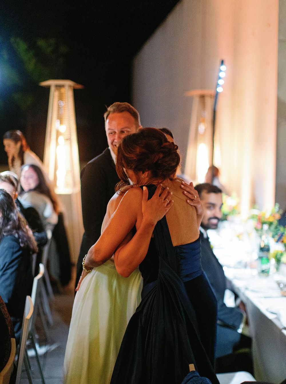 A bride and her mom embrace lovingly at wedding reception, surrounded by friends and family.