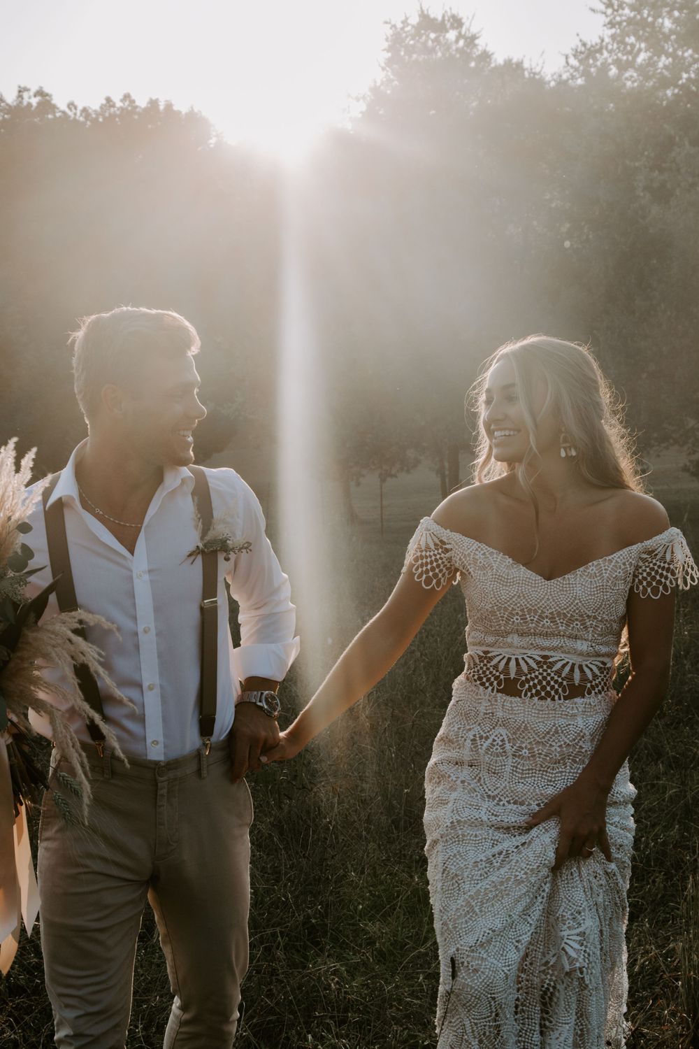couple holding hands and looking at each other, walking through a field with sun flares