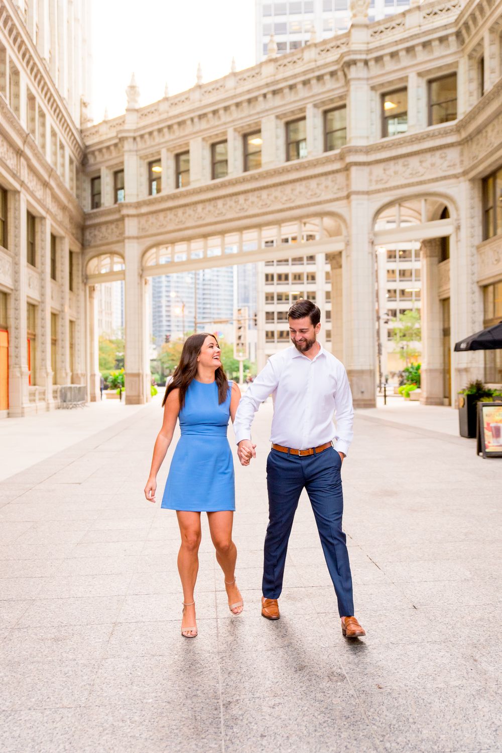 woman with brown hair in a light blue dress holding hands with fiance in front of the wrigley building in chicago
