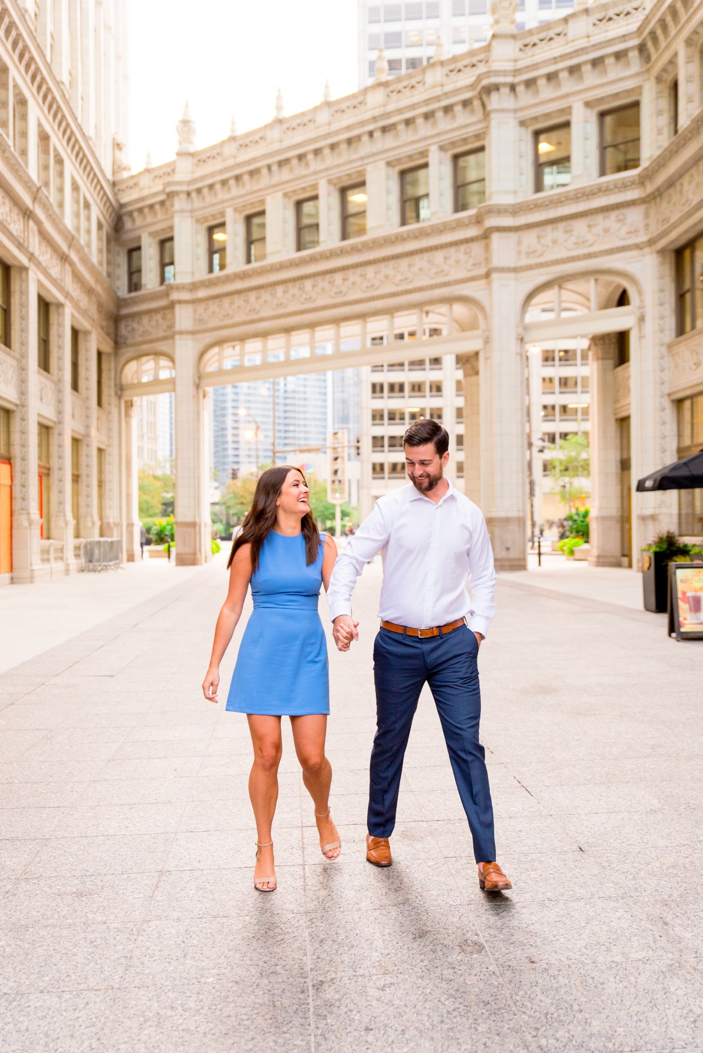 woman with brown hair in a light blue dress holding hands with fiance in front of the wrigley building in chicago