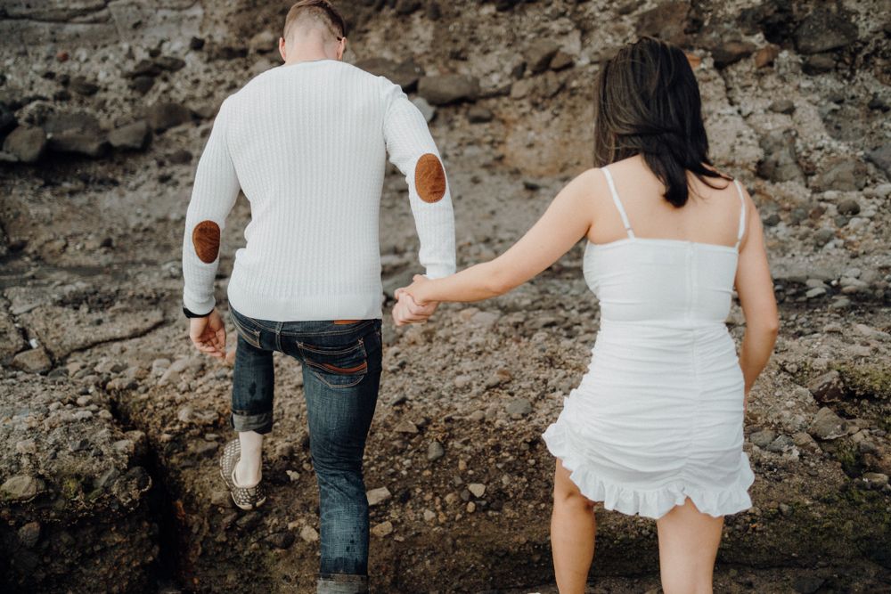 A couple in casual attire descends a rocky coastal slope near the ocean.