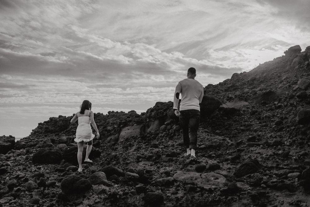 Two people traverse dramatic volcanic rocks along a black sand beach under cloudy skies.