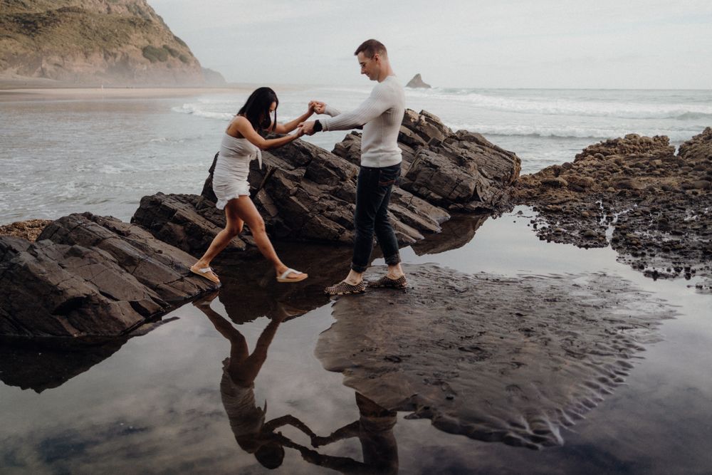 Two people carefully navigate rocky coastal formations as waves lap at the shore.