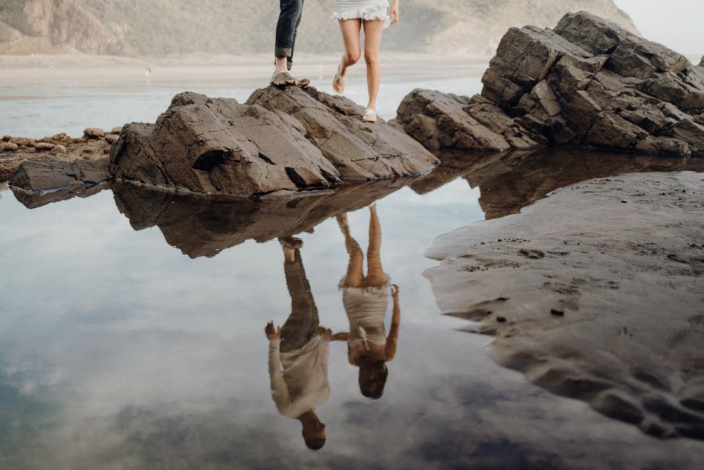 A couple stands on rocky coastal formations with their reflection perfectly mirrored in still water.