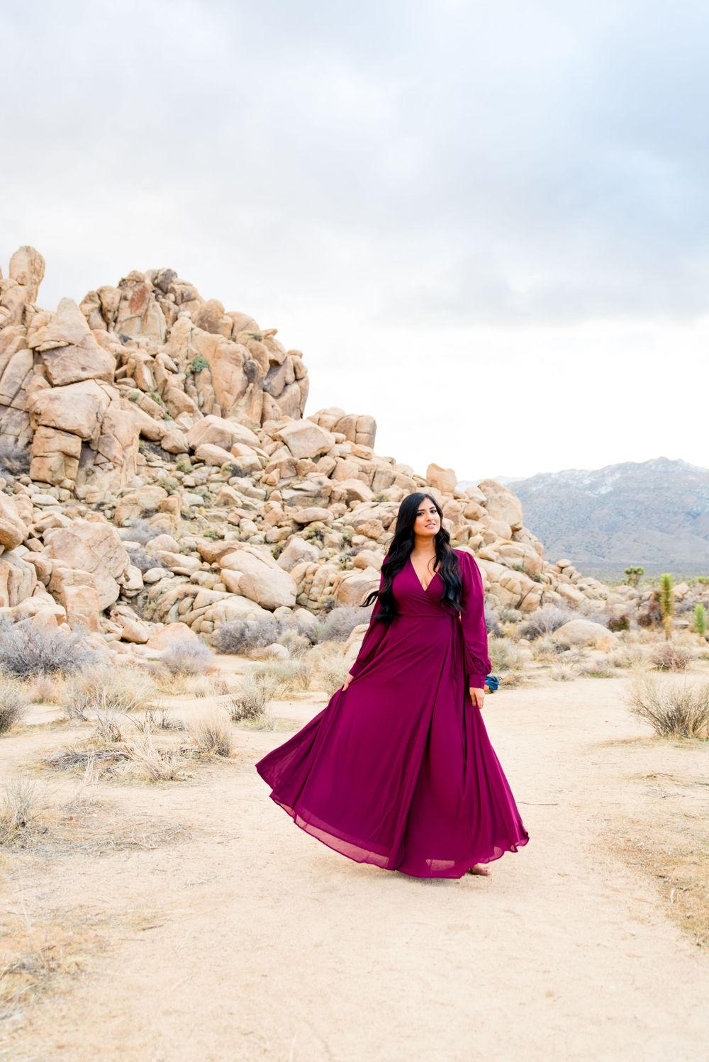 woman with long dark hair in red maxi dress looks to the side in front of boulders at Joshua Tree National Park