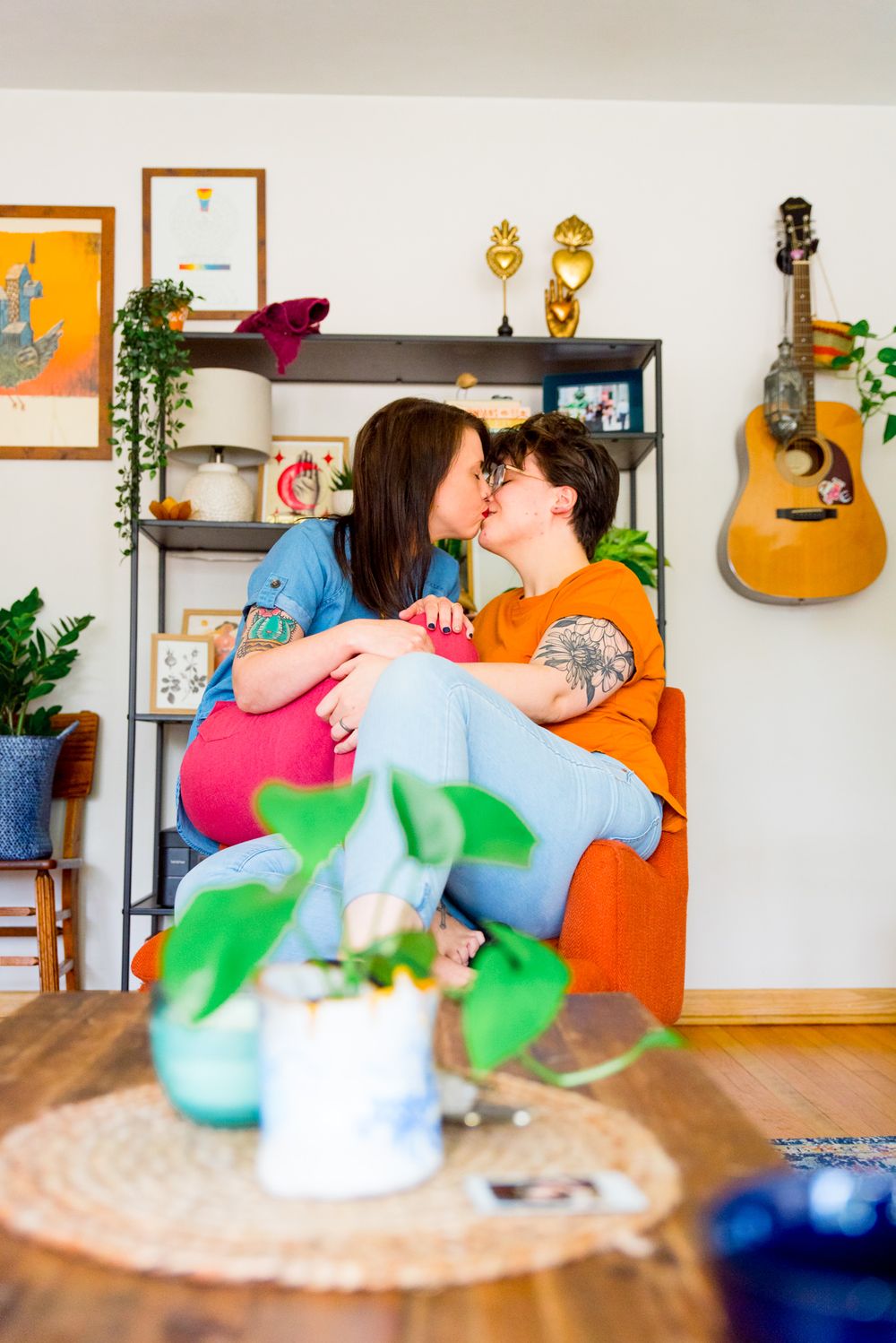 two woman in colorful clothing and colorful apartment full of plants sitting on an orange chair, kissing