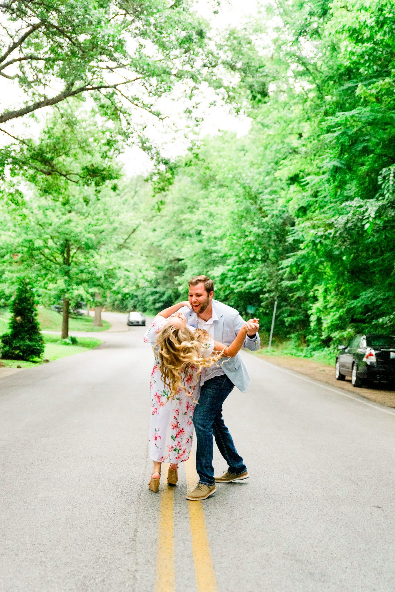 engaged couples in floral dress and light blue suit jacket and jeans dancing in middle of road under trees